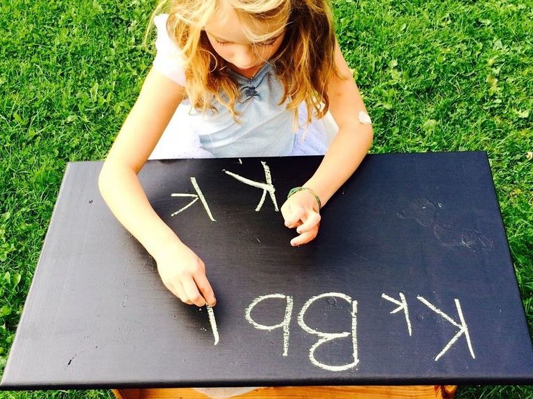 ein kleines Mädchen schreibet mit Kreide an einer Holztafel auf der Wiese