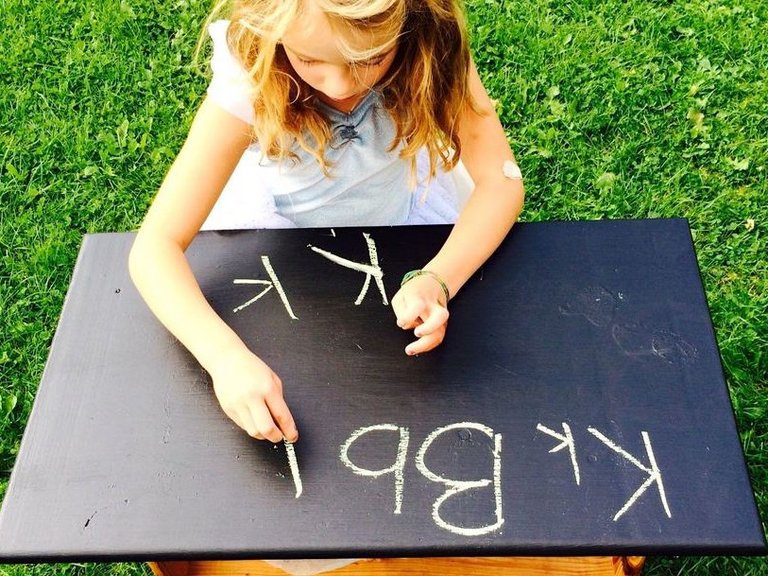 ein kleines Mädchen schreibet mit Kreide an einer Holztafel auf der Wiese