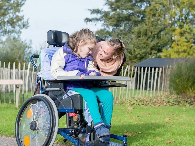 ein Mädchen im Rollstuhl - eine Betreuerin kümmert sich um sie und lächelt dabei