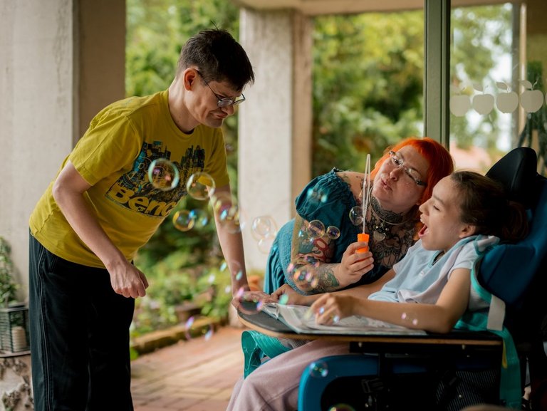 Zwei Jugendliche mit Beeinträchtigung und eine Mitarbeitende genießen den Moment, während bunte Seifenblasen um sie herum schweben.“